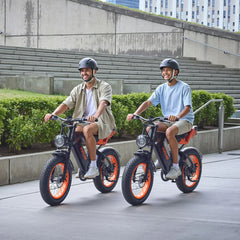 Two people riding VIPCOO VM6 Electric City Bikes on a city street with buildings and greenery in the background. - Pogo Cycles, UK