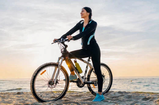 Woman riding a silver fat tyre electric bike on a sandy beach at sunset, side view.