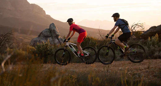 Two cyclists riding electric bikes on a mountain trail at sunset, side view